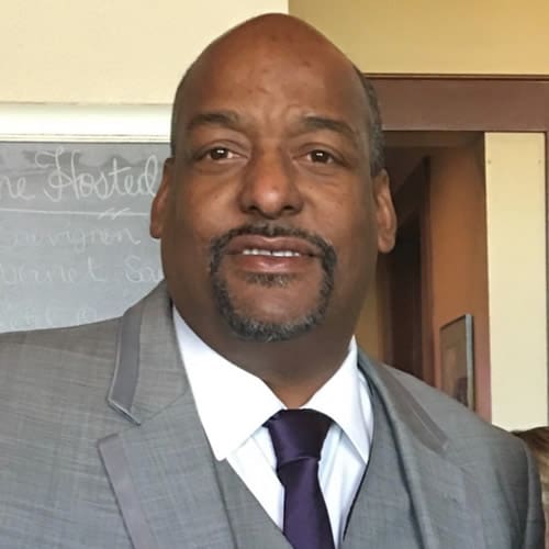 A man in a gray suit and purple tie smiling indoors.